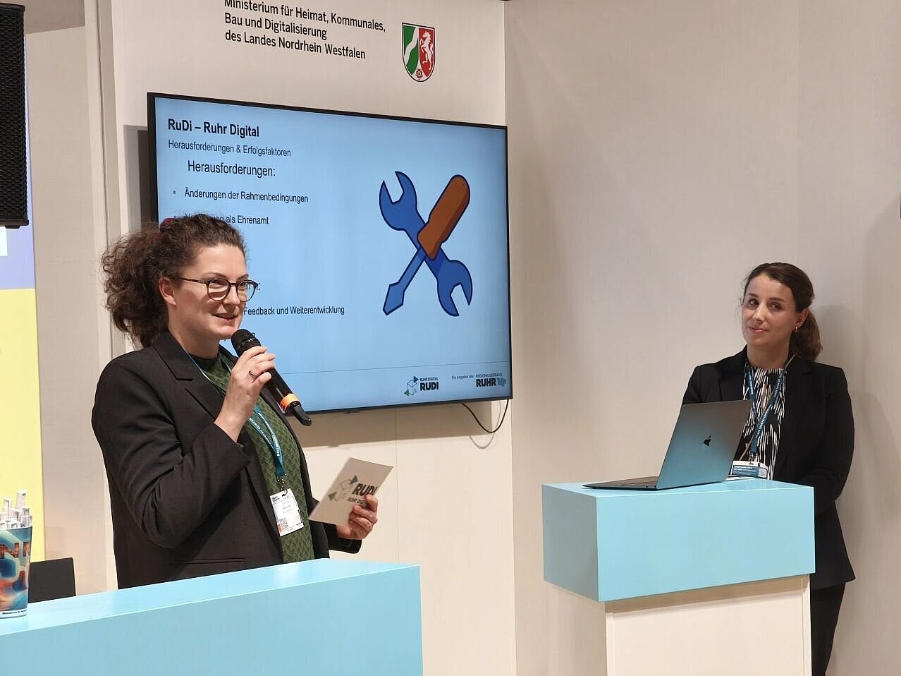 Zwei Personen an Rednerpulten, eine Person hält ein Mikrofon und Moderationskarten. Hinter ihnen ein Bildschirm, auf der eine Präsentation läuft. 