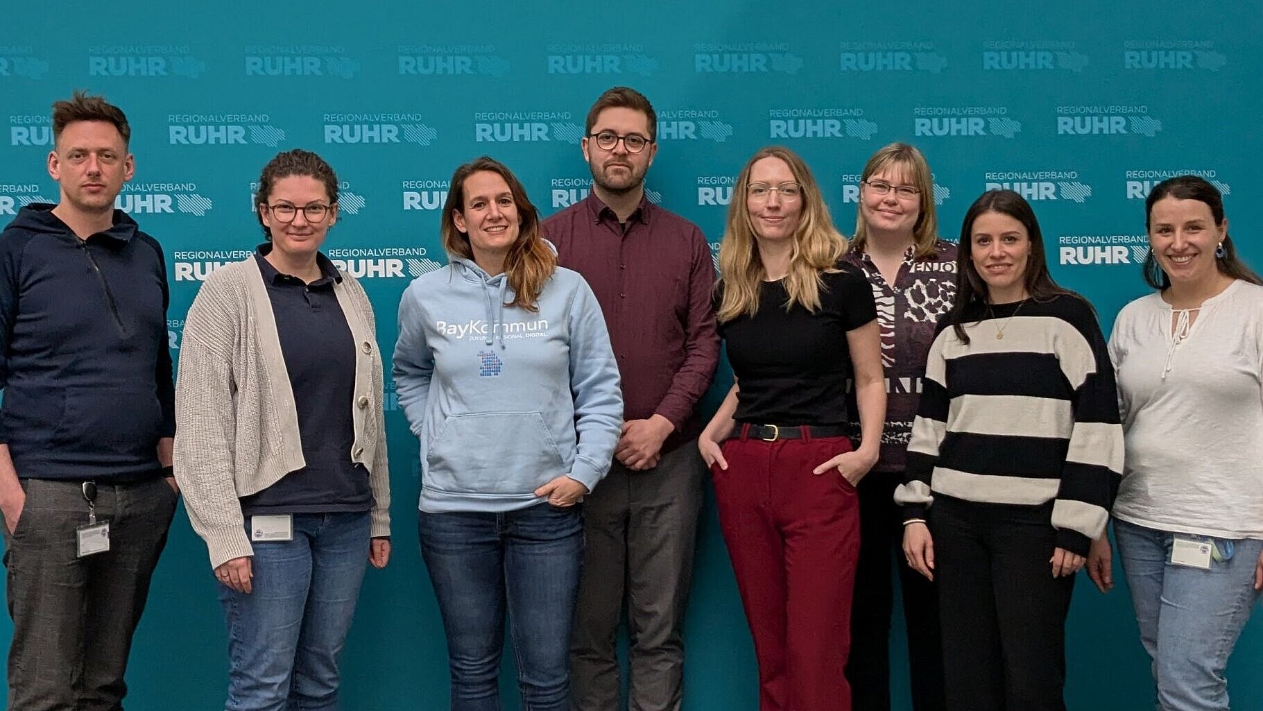 8 Personen nebeneinander vor einer petrolfarbenen Fotowand. 